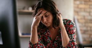 Woman at desk holding her head, showing how stress sabotages your sleep