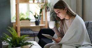 Woman wrapped in a blanket holding a white mug and reading a tablet as part of a night routine that actually works in a calming bedroom environment for sleep