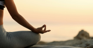 Person sitting cross-legged on a yoga mat near the water at sunset, practicing a proven breathing technique with hand in a meditative position.